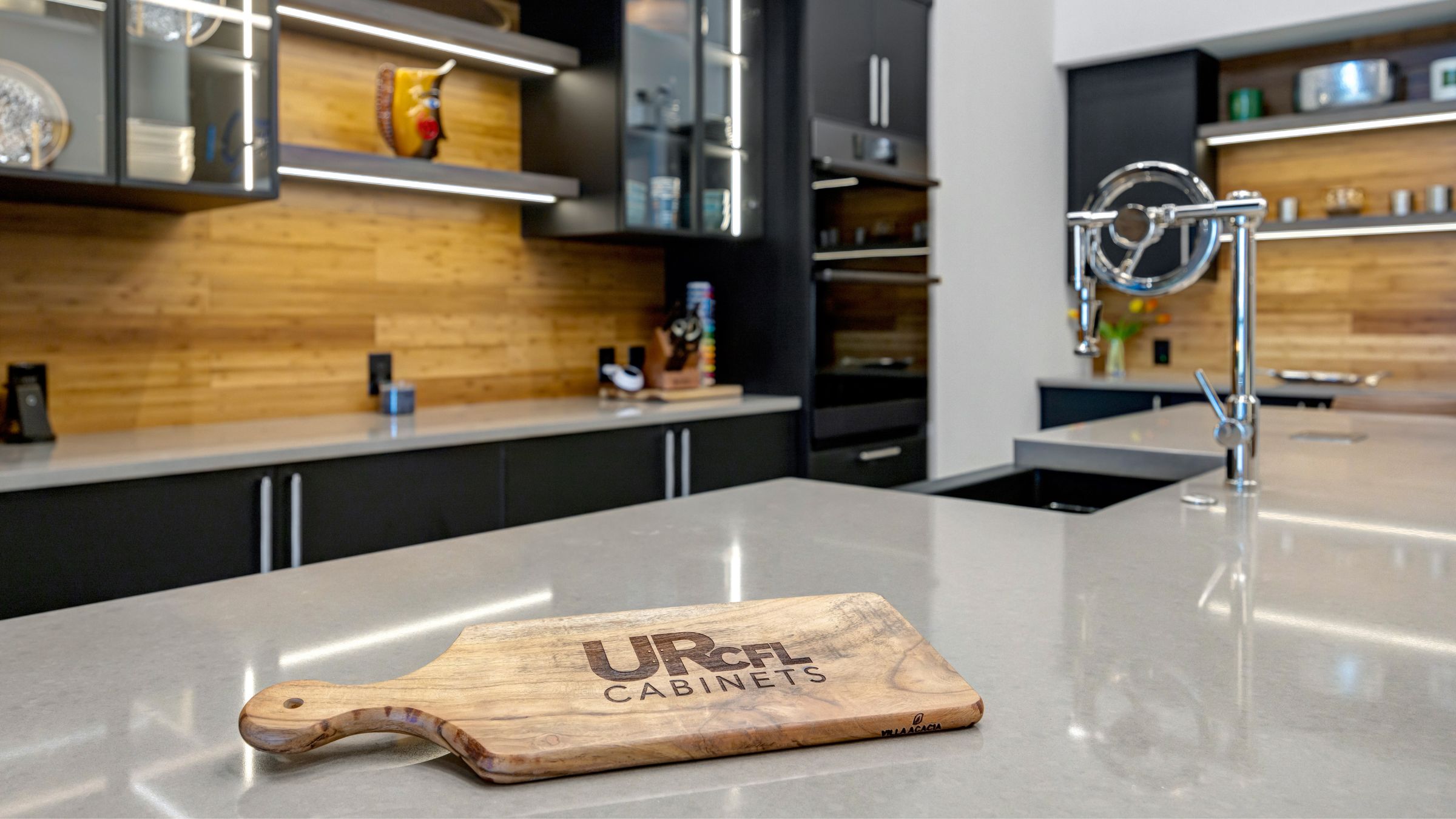 A modern kitchen featuring white shaker-style upper and lower cabinets with sleek silver handles. The central island is a dark charcoal color with a smooth white countertop, providing seating for three with wooden bar stools. Stainless steel appliances, including a built-in oven and microwave, are integrated into the cabinetry. Pendant lights hang above the island, and recessed lighting brightens the entire space. The flooring is a light wood tone, adding warmth and contrast to the clean, contemporary design.
