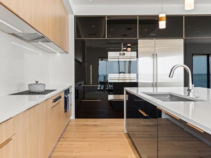 A contemporary kitchen featuring a combination of white and beige cabinets paired with black accents. The large central island has a white marble countertop with subtle gray veining and includes built-in storage. Above the island hang two sleek black pendant lights. Stainless steel appliances are integrated into the cabinetry, and the backsplash is a light neutral color. The overall design balances warmth and contrast, creating an inviting and stylish space.